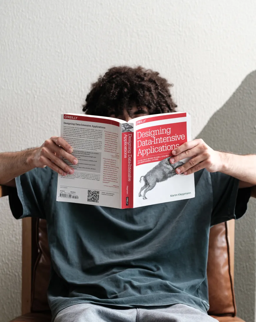 A curly-haired person in a dark grey t-shirt, holding a book called “Designing Data-Intensive Applications” by Martin Kleppmann.