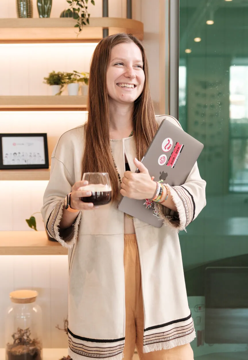 A blond woman, smiling and holding a space grey MacBook pro covered with Atolye15 stickers and a glass of coffee.