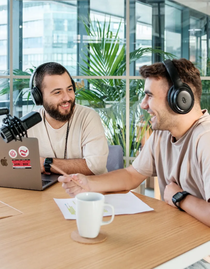 Two male Atolye15 employees with headphones on, sitting at a table, smiling, and talking to a podcast microphone.