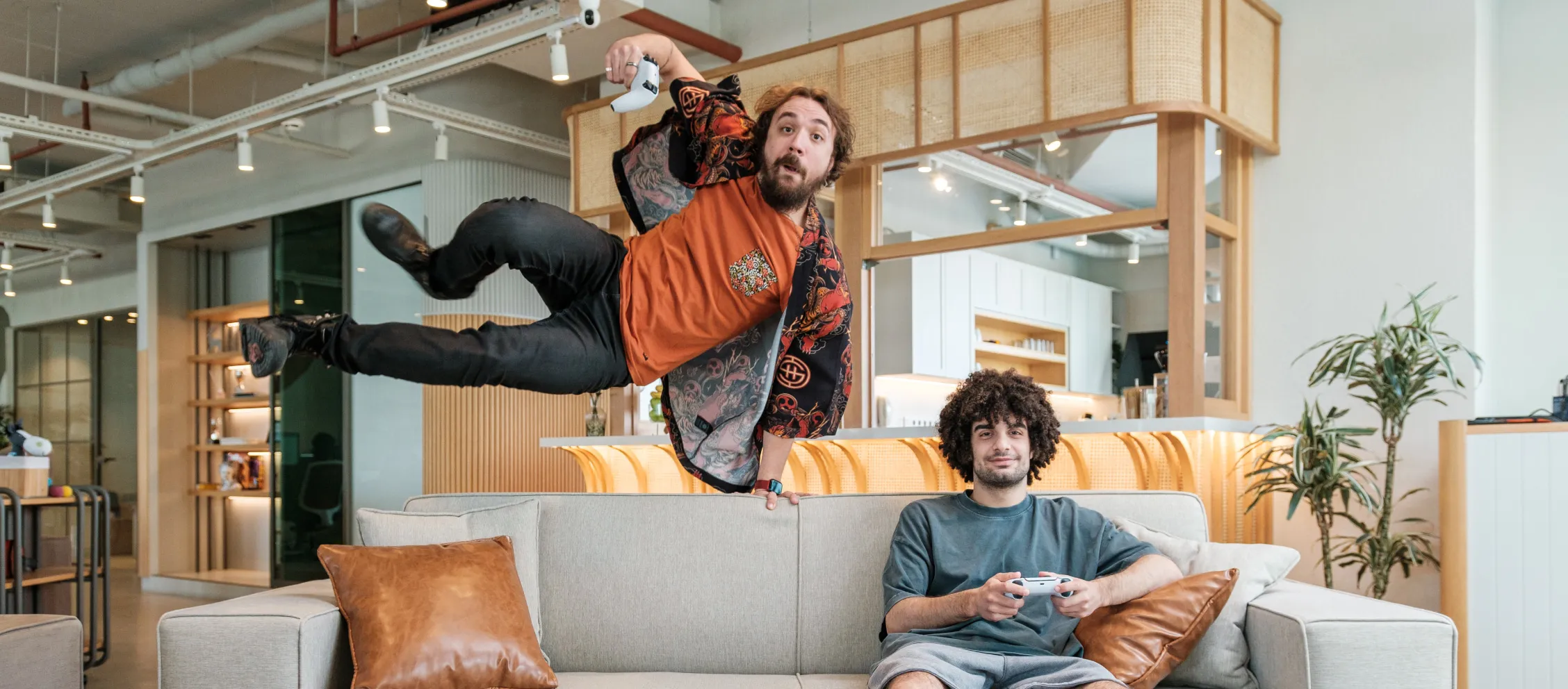 A man in an orange shirt jumping onto a couch where a curly-haired man is sitting down with a game console in his hands.