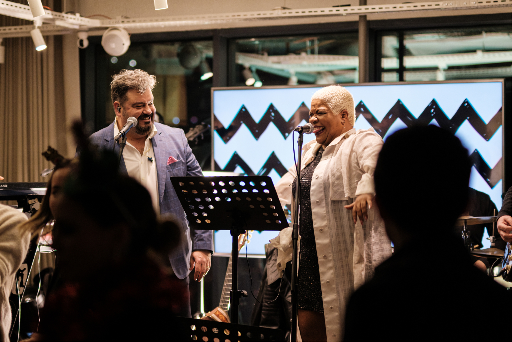 A bearded man in a blue suit and a short-haired woman in a white shirt singing into microphones on a stage.
