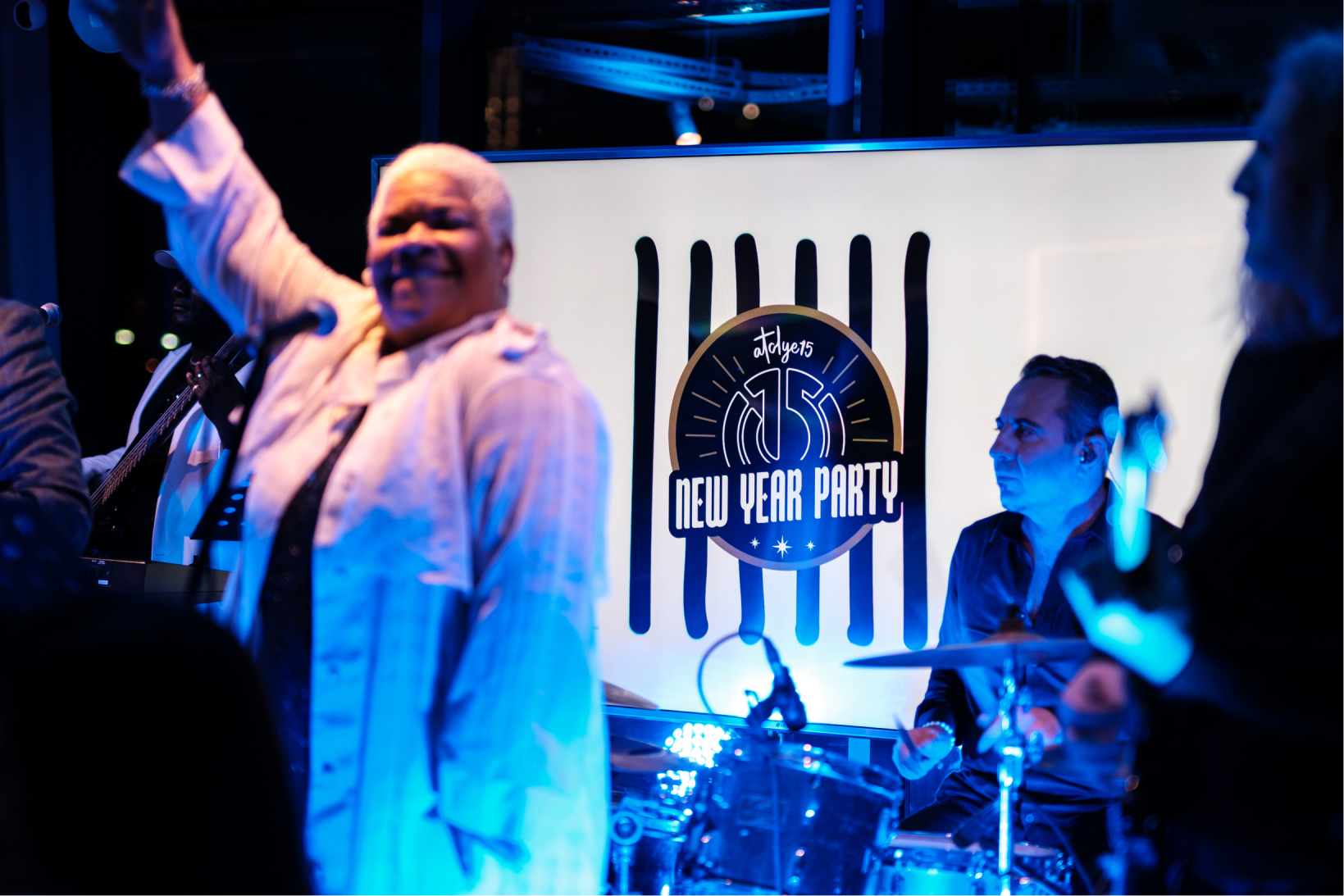A drummer and a female singer with short blonde hair in front of a screen that says “Atolye15 New Year Party.”