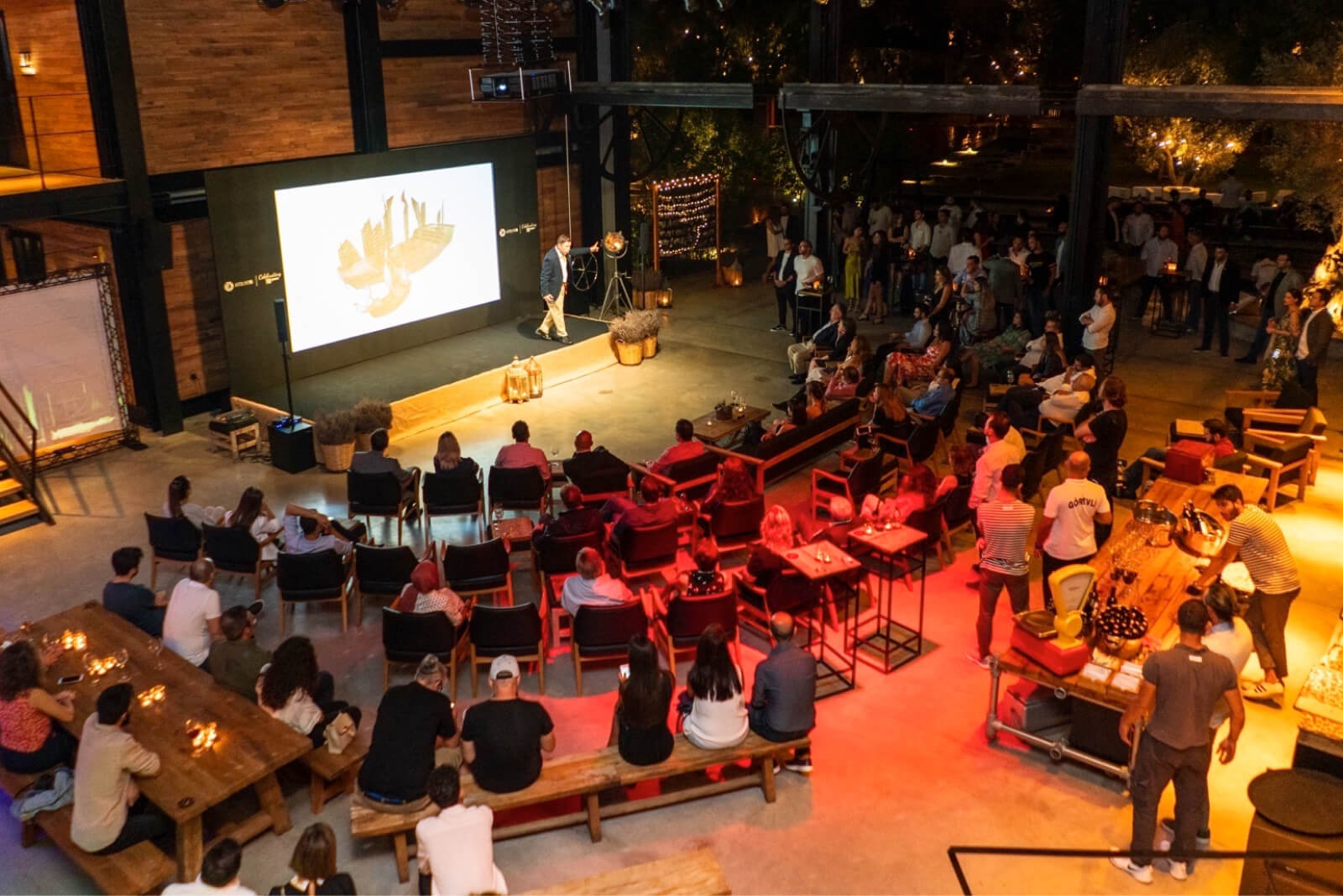A group of people sitting in chairs, looking at a screen, and listening to a presentation at Atolye15's 10th year party.