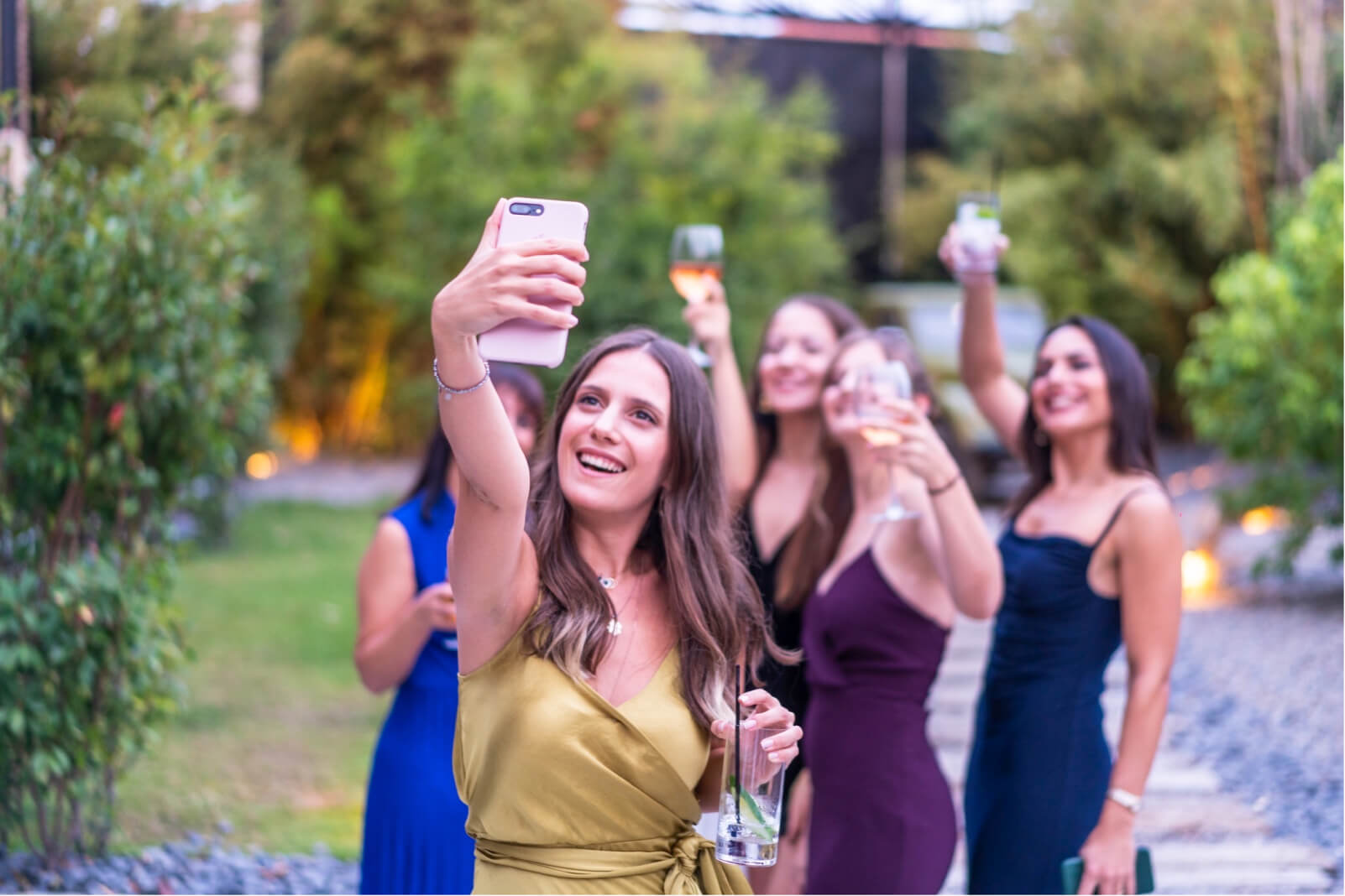 A group of young women in evening dresses raising their glasses in the air and taking a selfie in a green garden.
