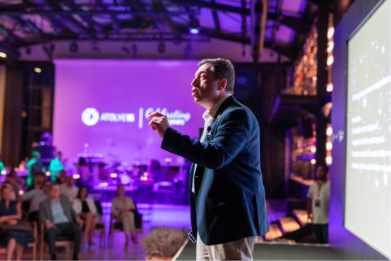 A middle-aged man in a blue jacket giving a speech on a stage in front of a screen that says “Atolye15 Celebrating 10 years.”
