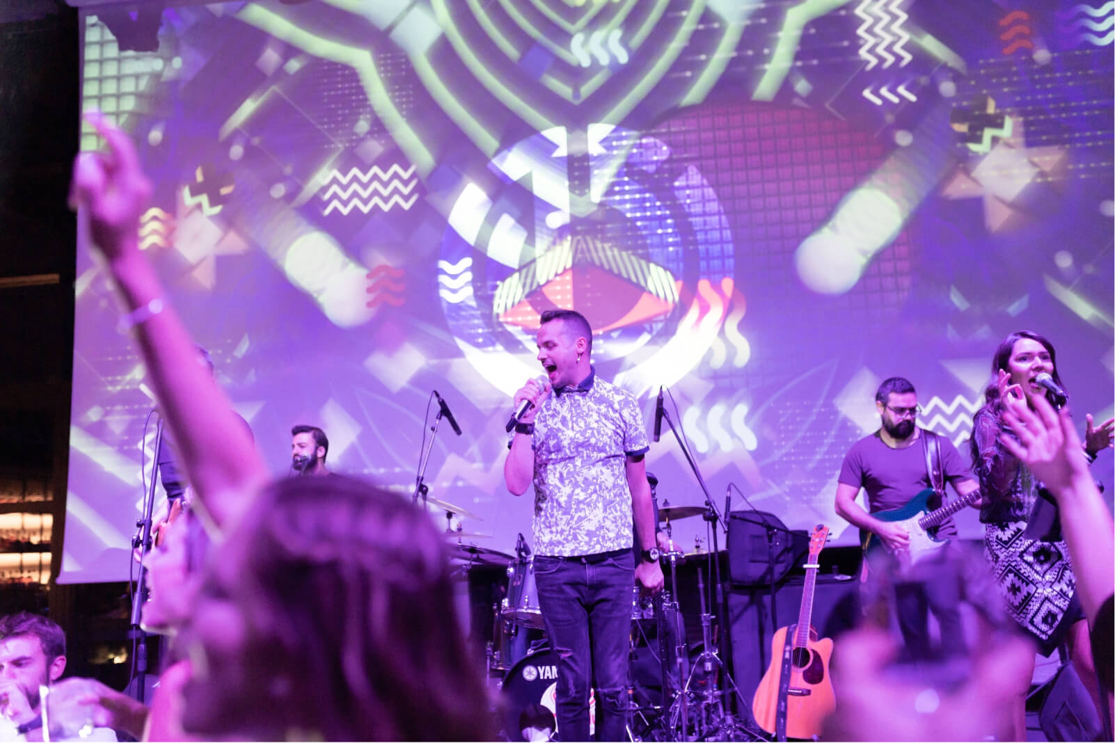 A male singer and his music band, singing in front of the Atolye15 logo while people are dancing with their hands in the air.