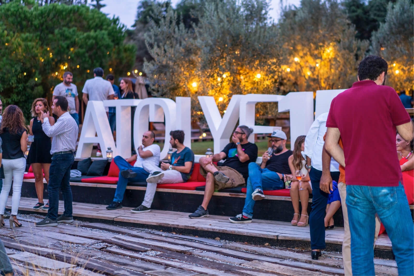 People talking and sitting in front of an Atolye15 letter statue among trees with yellow string lights.
