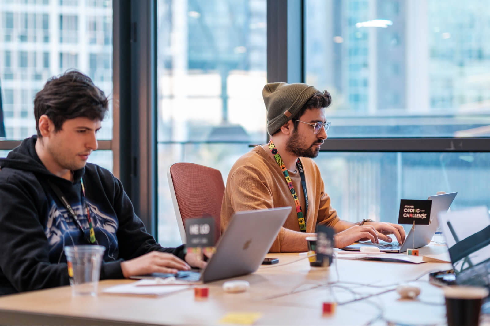 A man in a black hoodie and a man with a hat sitting at a desk working on their laptops and a card saying Coding Challenge.