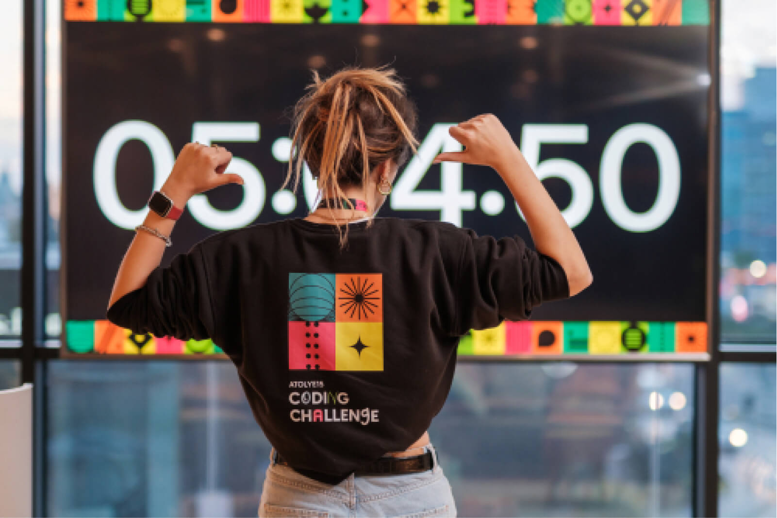 The back of a blond girl with her hands pointing at the Atolye15 Coding Challenge logo at the back of her black sweatshirt.