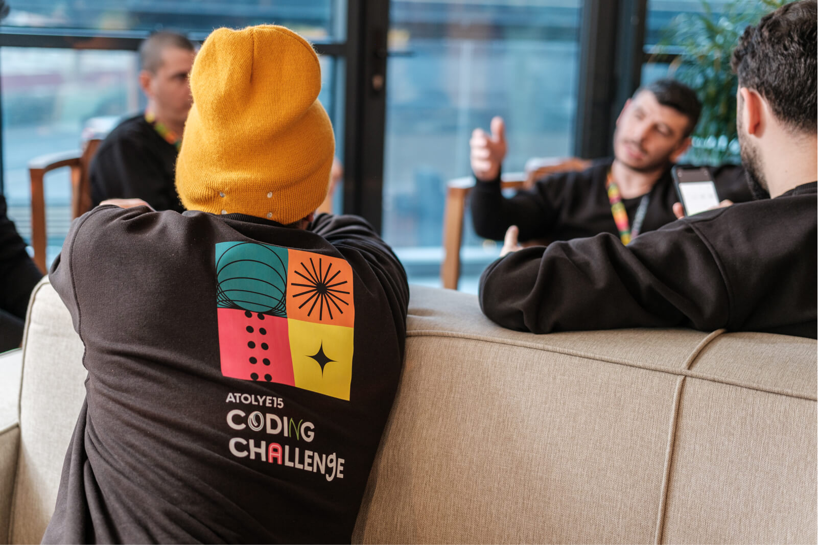 A man wearing a yellow hat and a black sweatshirt that has the Atolye15 Coding challenge logo on the back.