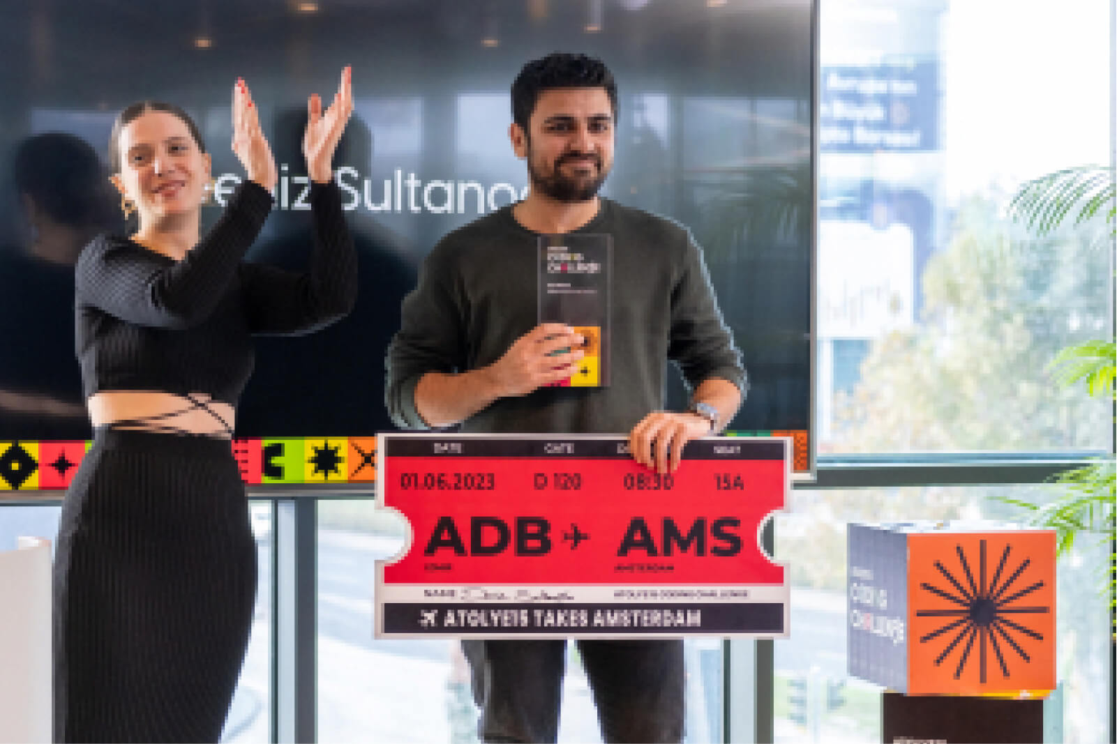 A man holding the Atolye15 Coding Challenge winner's cup and a plane ticket award to Amsterdam and a woman clapping on stage.