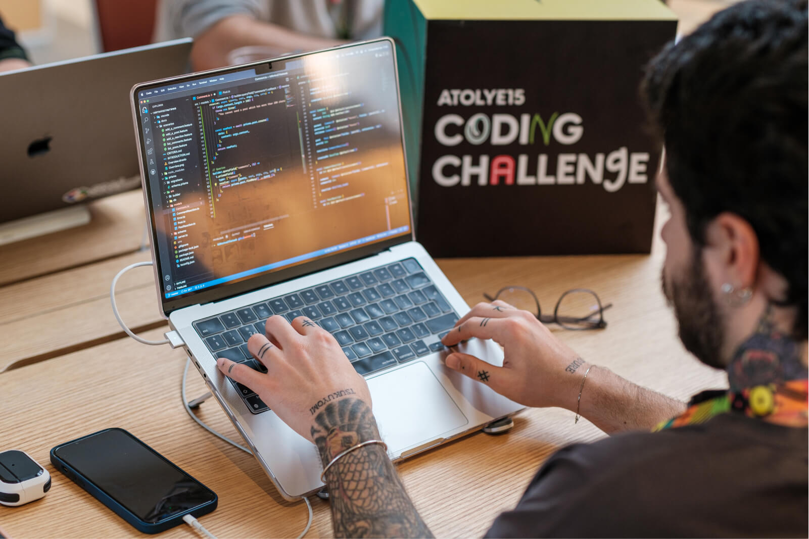 A man with tattoos coding at the wooden desk which has a laptop, an iPhone, glasses, and an Atolye15 Coding Challenge cube.