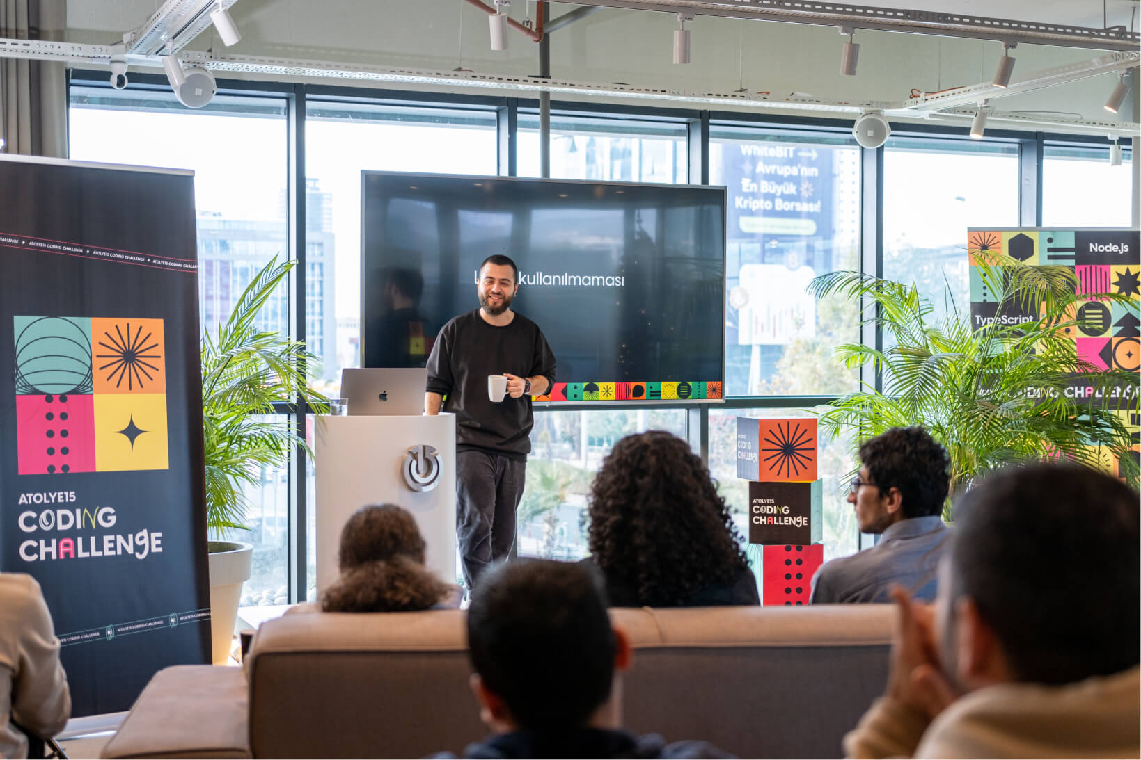 A man in a black sweatshirt holding a white mug, talking to people on a stage, and an Atolye15 Coding Challenge banner.