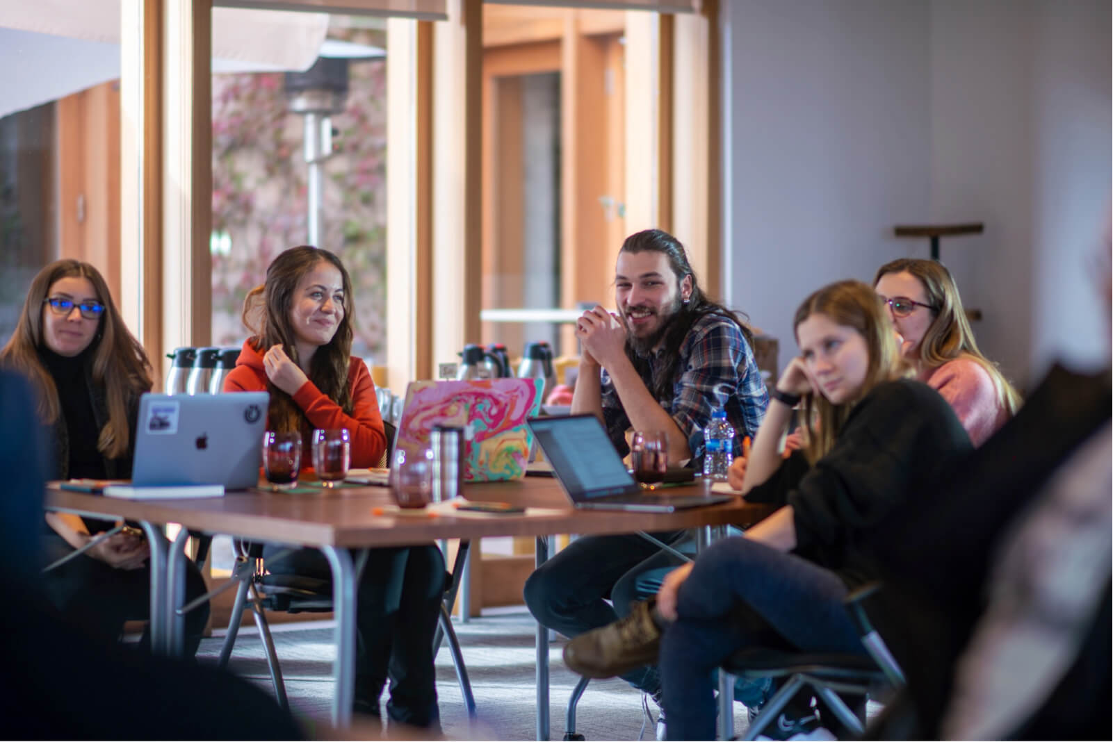 Four women and a long-haired man sitting around a table in front of MacBooks, smiling and having a meeting.