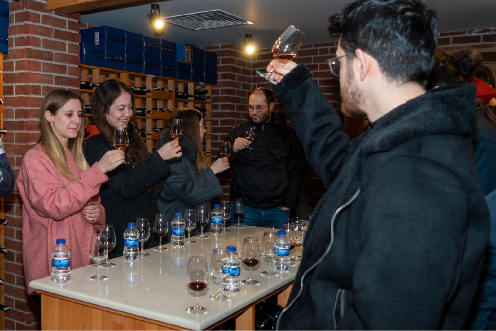 People standing around a marble countertop with water bottles and glasses, tasting wine in a brick-walled wine cellar.