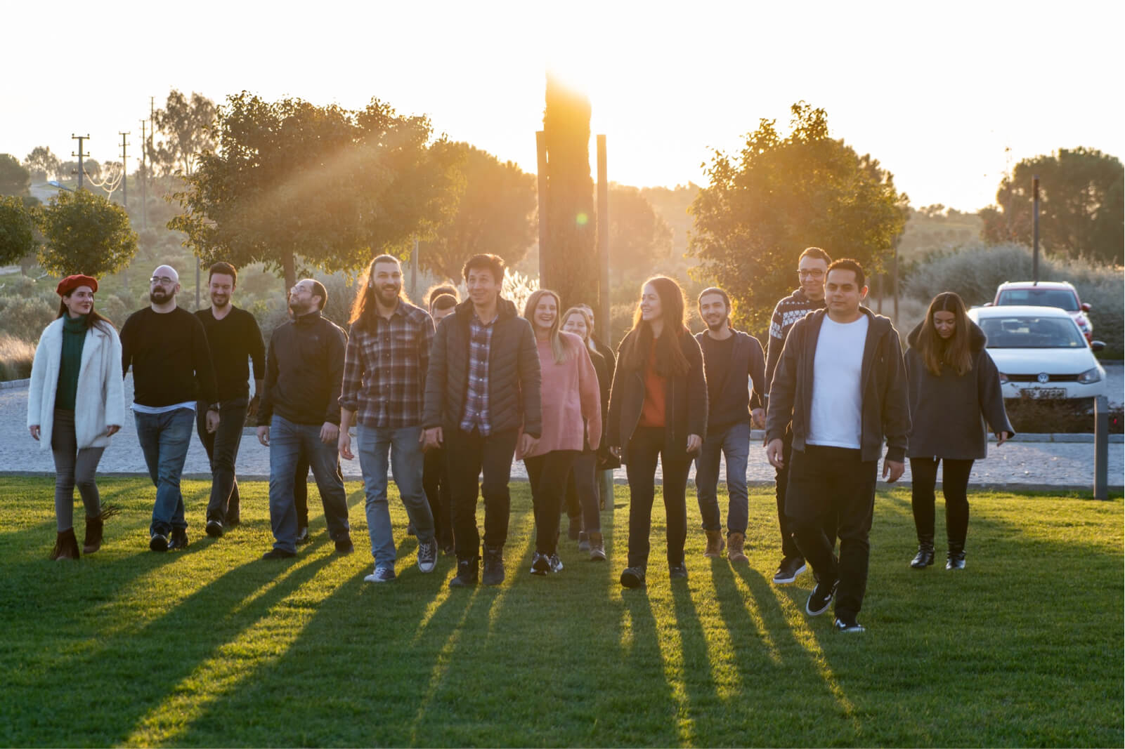 A group of people smiling and walking on green grass with sunlight, trees, and cars behind them.