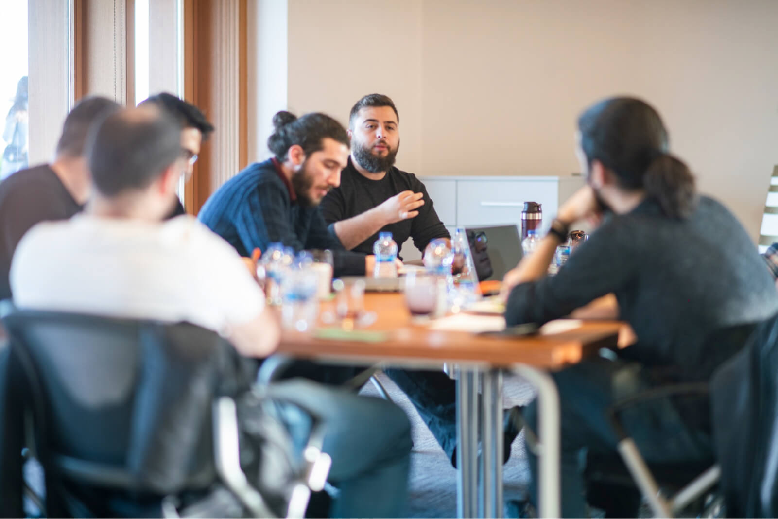 A group of men sitting around a wooden table filled with glasses, water bottles, and laptops, having a meeting.