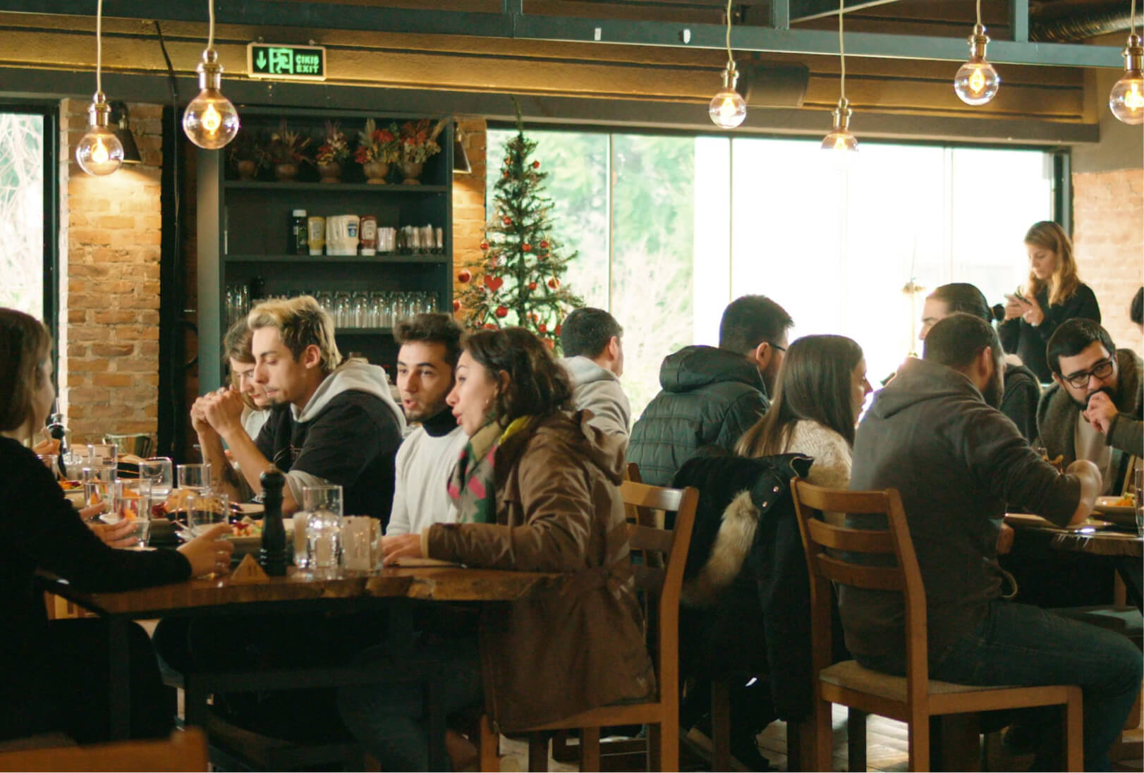People sitting at tables in a restaurant eating food and having conversations next to a Christmas tree under light bulbs.