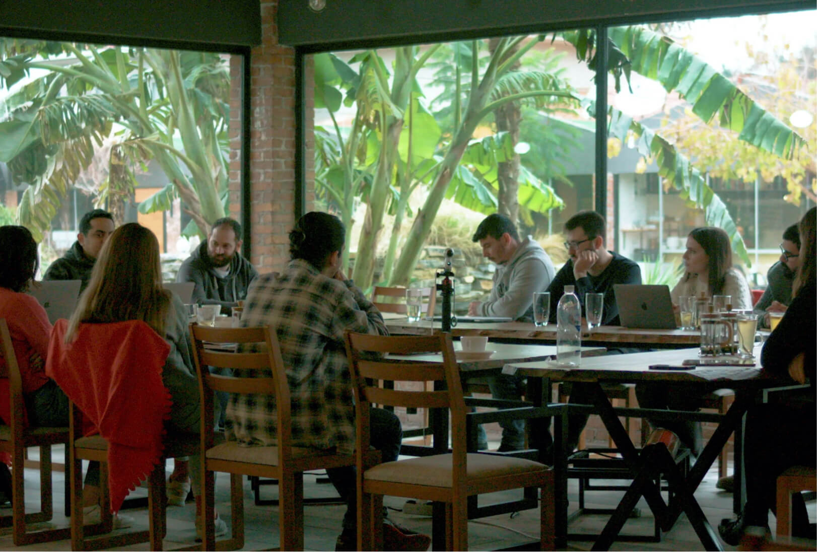 People sitting around a dinner table with glasses and water bottles and working on laptops at a restaurant by palm trees.