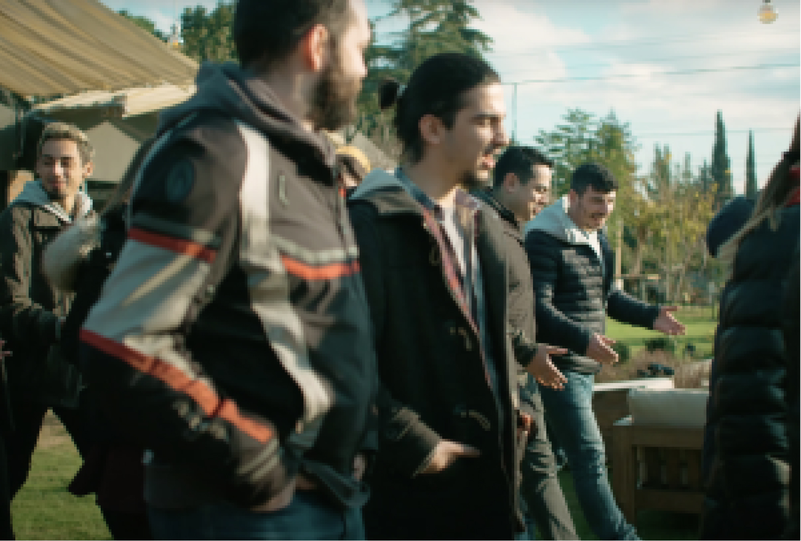 A group of men in jackets walking on the grass next to trees while having a conversation with each other.