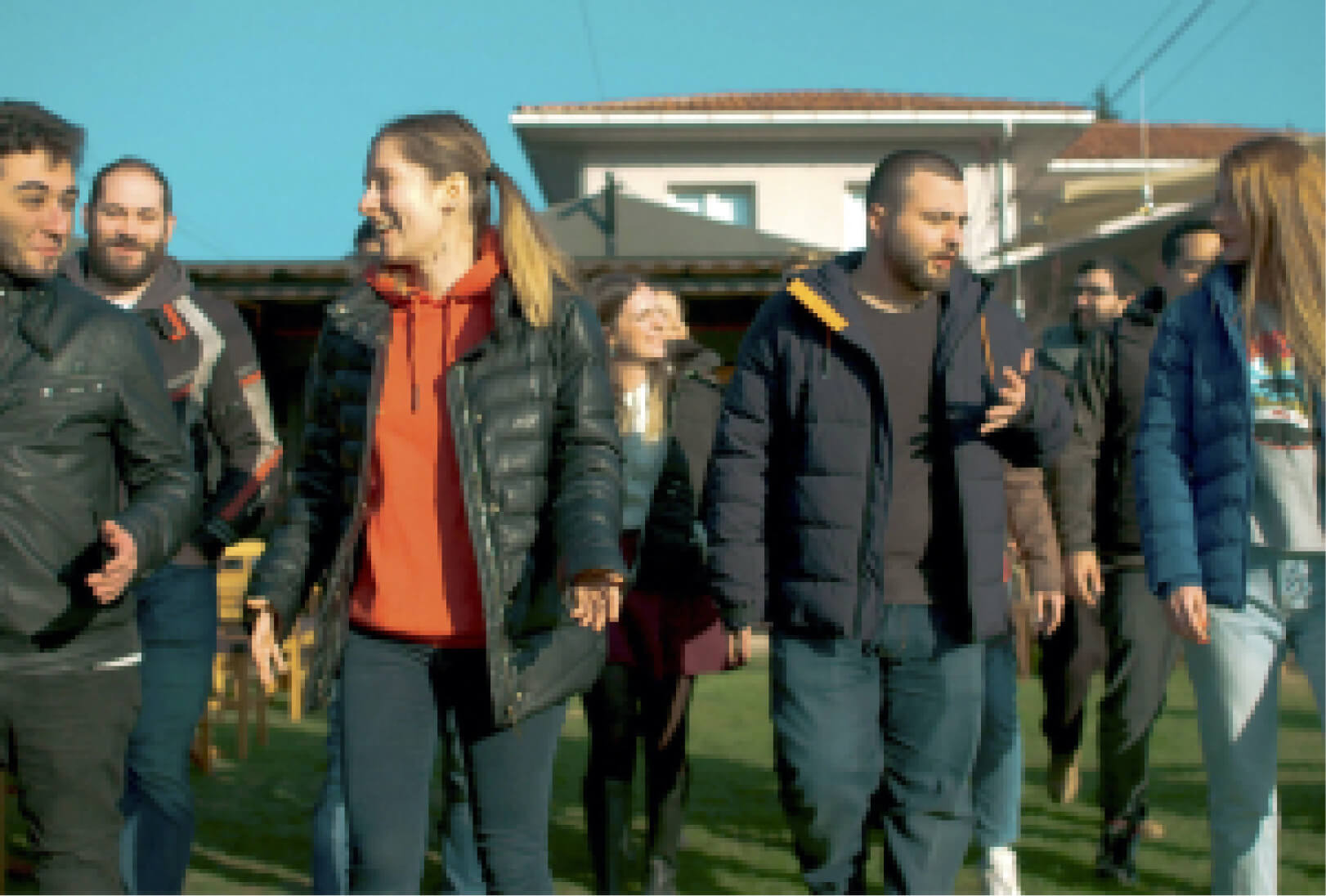 A group of people in puffer jackets, walking on the grass while having a conversation in front of a villa.