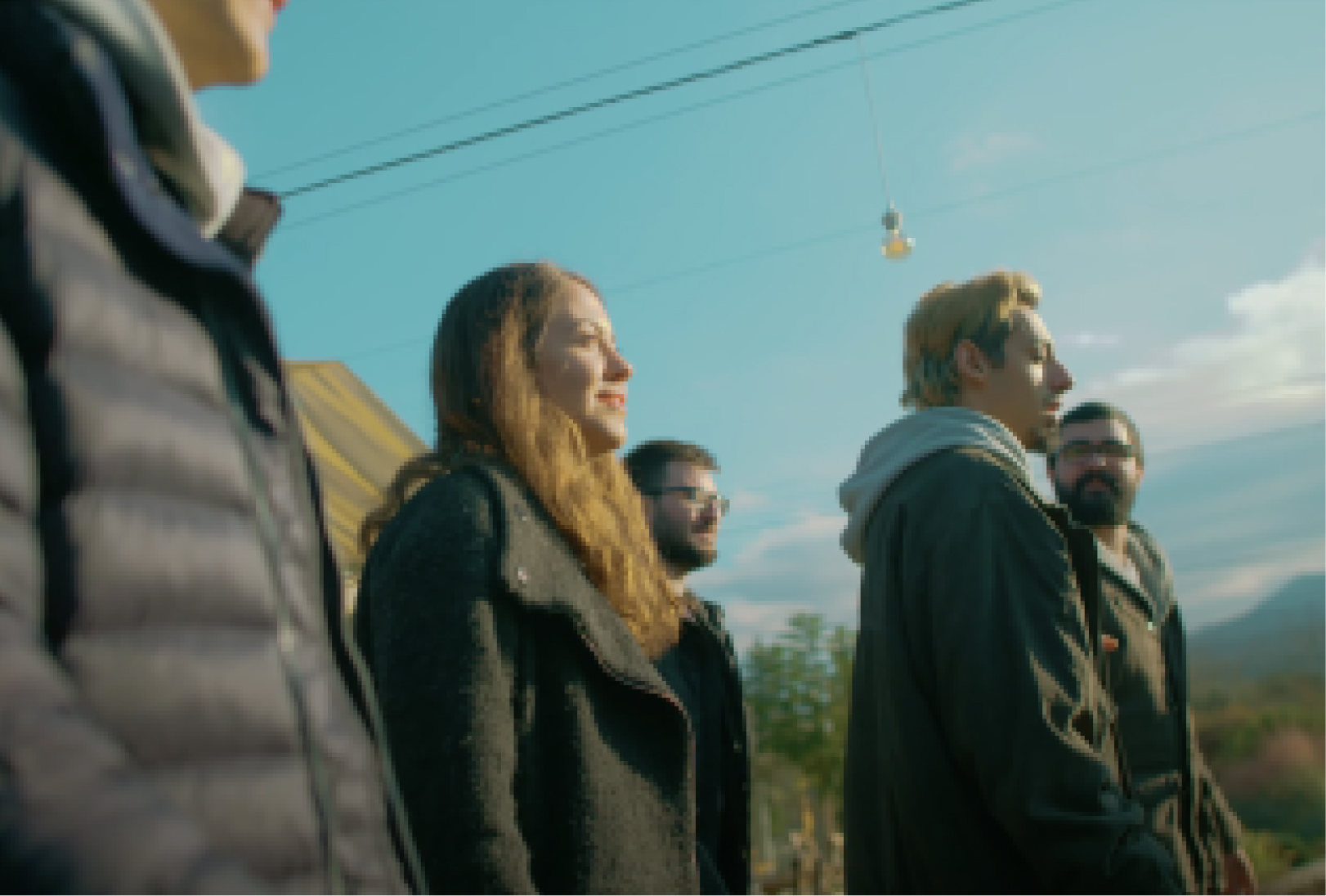 Two men wearing glasses, a blonde man, and a long-haired woman in a coat walking by trees under the clouds and a blue sky.