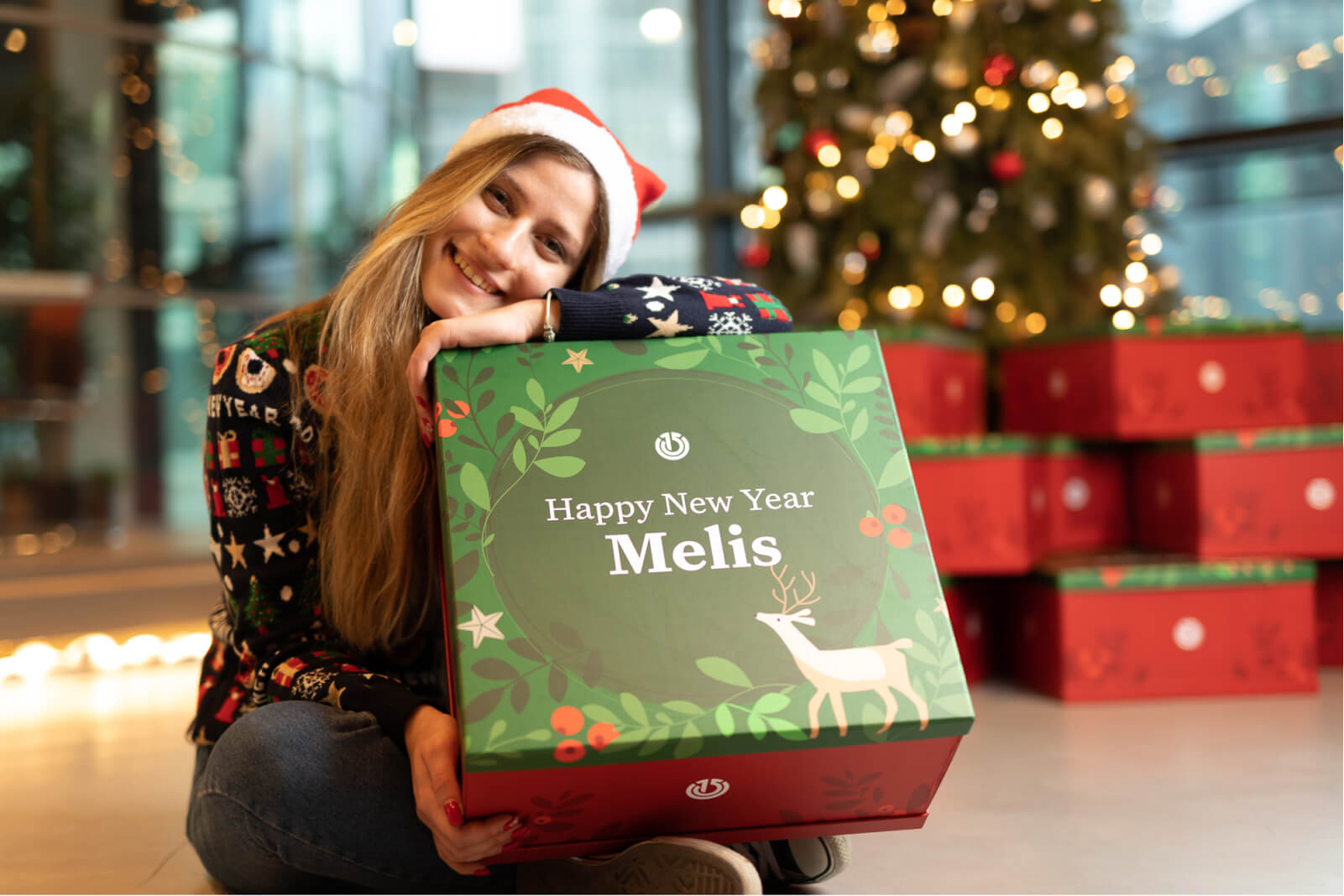 A blond girl in front of a Christmas tree, wearing a red Santa hat holding a gift box that says “Happy New Year Melis.”