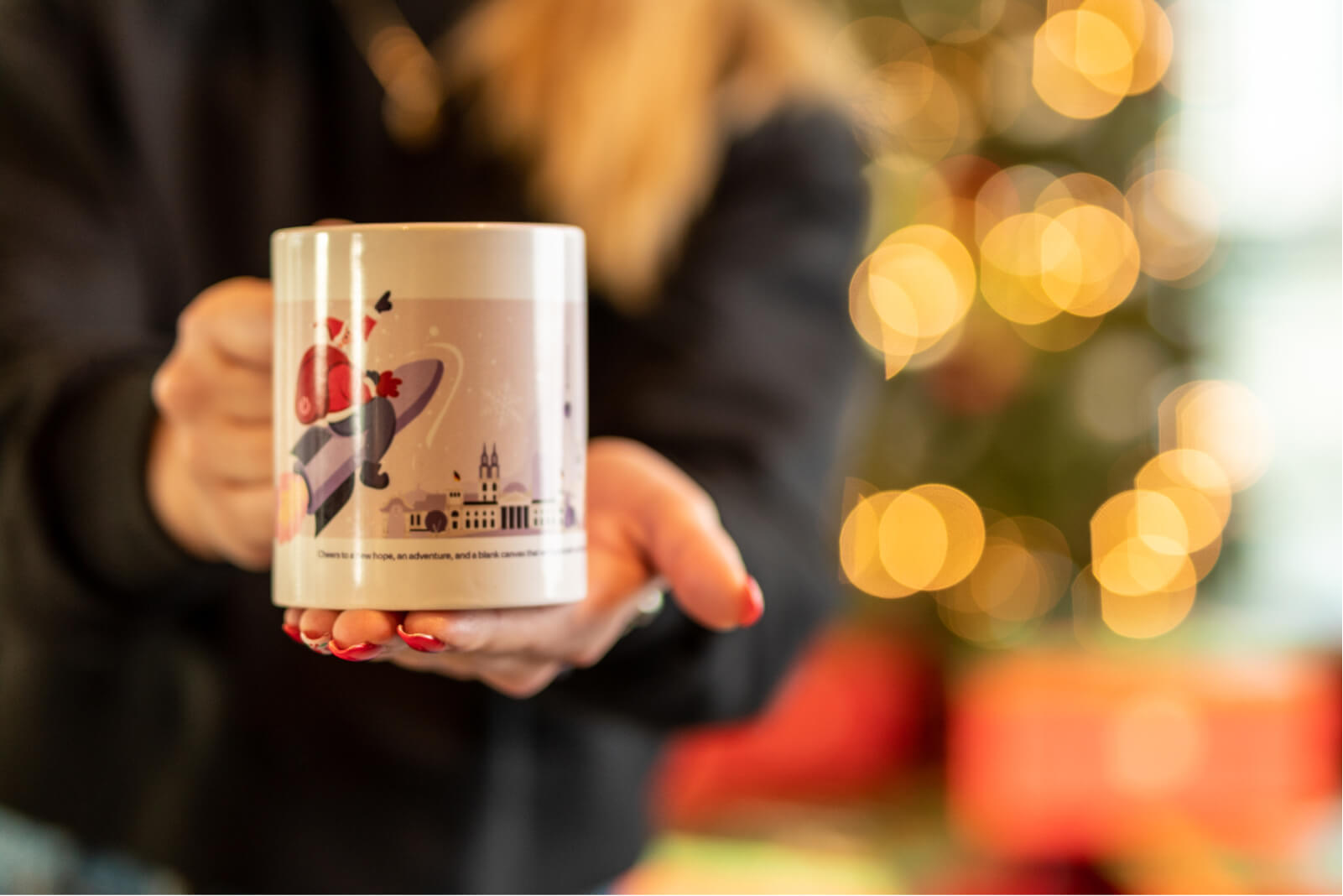 A pair of hands with red nails holding a Christmas coffee mug with an illustration of Santa flying over buildings on a rocket.