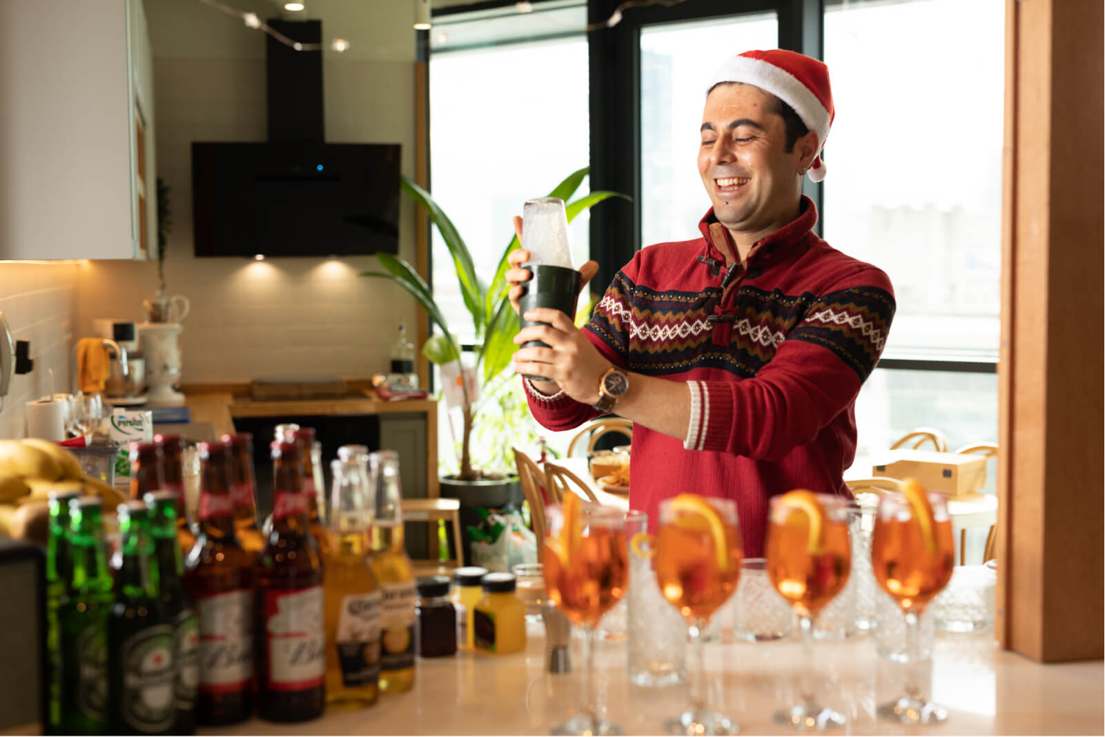 A smiling man wearing a Santa hat and a red Christmas sweater making cocktails by a kitchen counter with beer bottles.