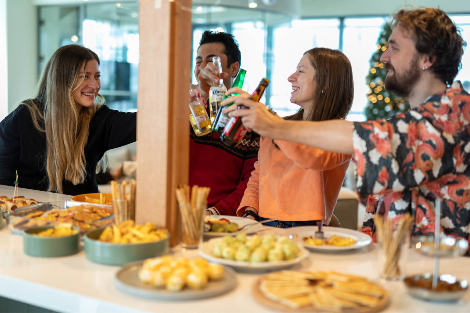 Two men and two women smiling and raising their beer bottles in front of a kitchen counter filled with plates of food.