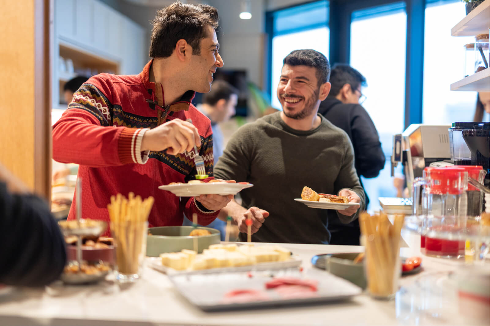 A man in a red sweater smiling at a man in a grey sweatshirt holding a plate and a fork in front of a counter filled with food.