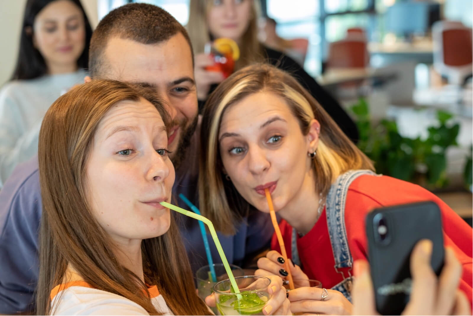 A smiling man between two blonde girls sipping their cocktails with a straw and posing for a selfie.