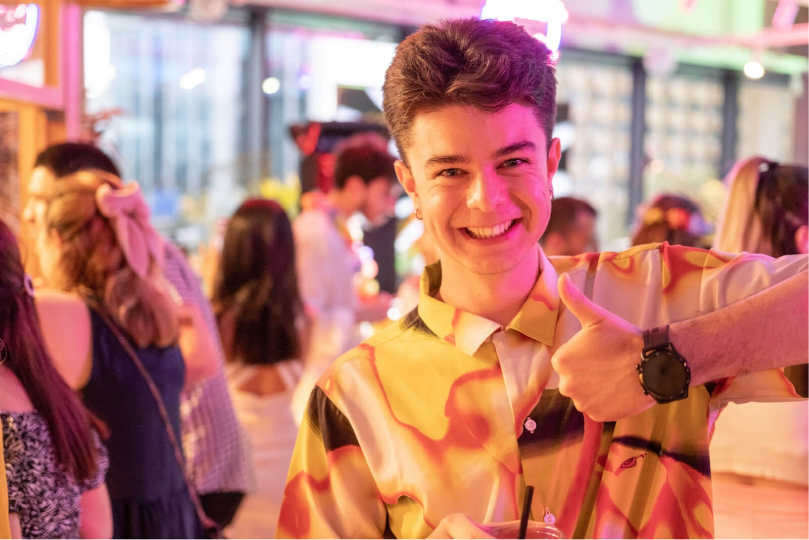 A young boy at a crowded party wearing a colorful shirt and a black watch, smiling, and giving a thumbs-up sign.