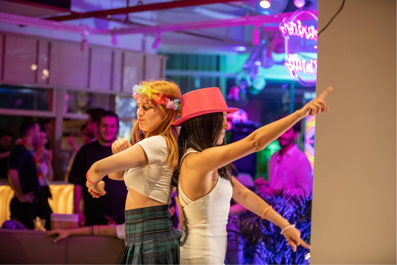 A blonde girl wearing a flowery headband and a green plaid skirt with her back against a brunette girl in a white dress and pink fedora.