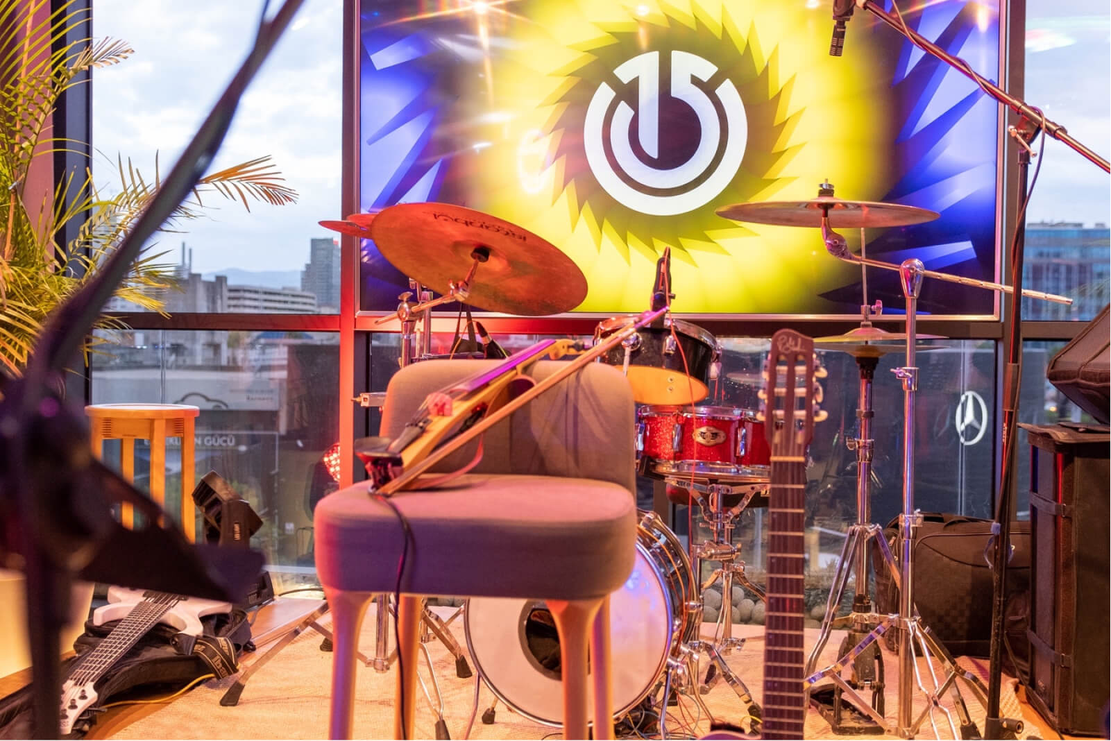 A grey chair, electric guitar, microphone, and drums on a stage and a screen that shows the Atolye15 logo.