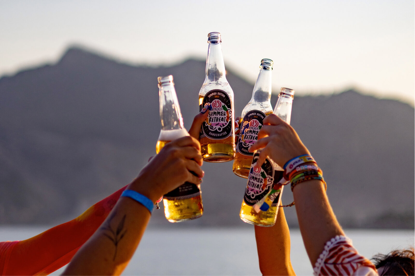 Hands in the air, raising custom-made Atolye15 Summer Retreat beer bottles with mountains and the sea.