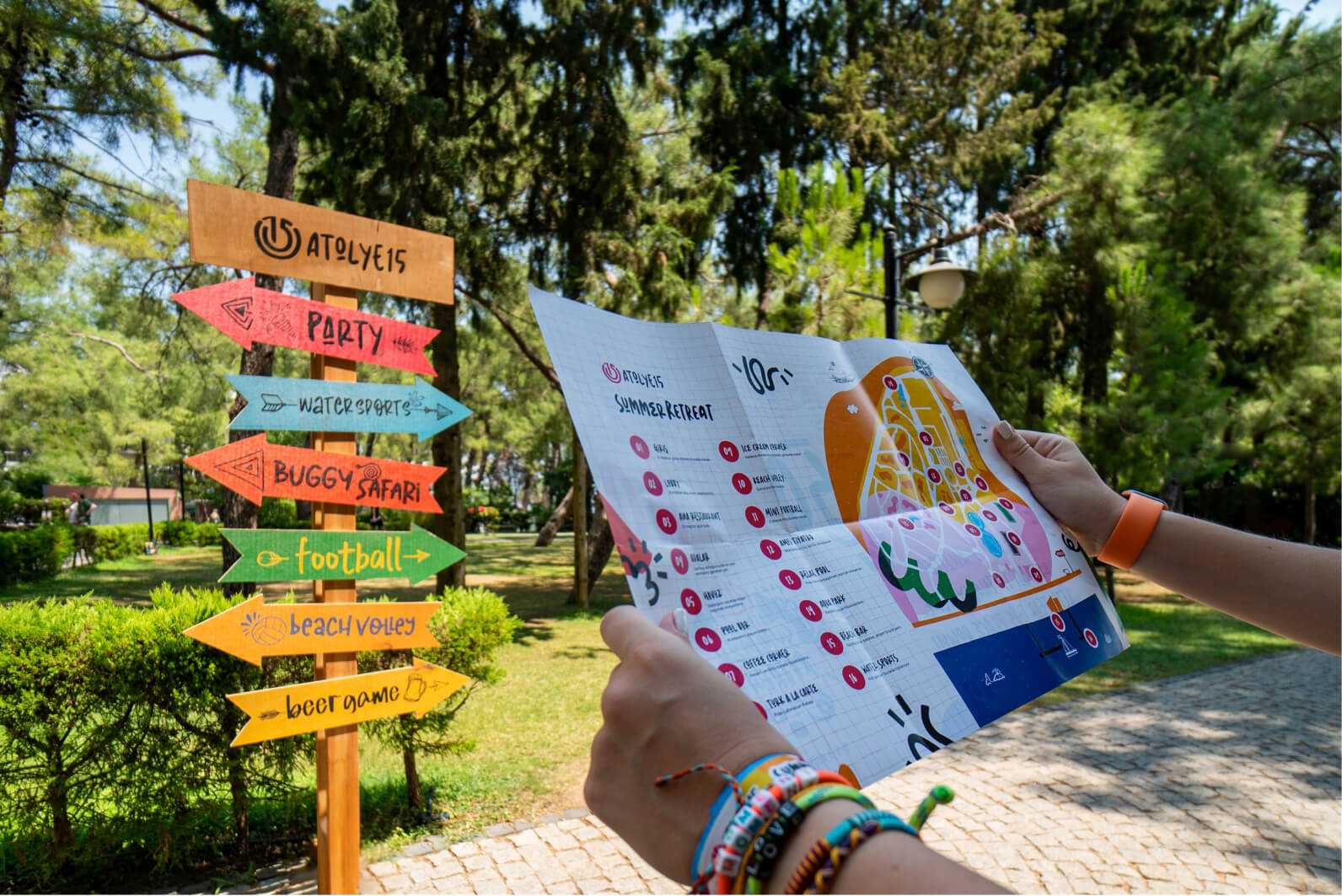 A pair of hands holding an Atolye15 Summer Retreat map in front of a colorful event sign with arrows pointing to the party and more.
