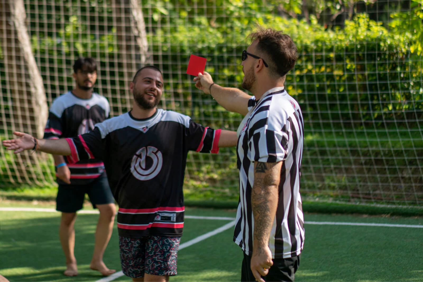 A man in summer shorts and a football uniform which has the Atolye15 logo on it and a referee showing him a red card.