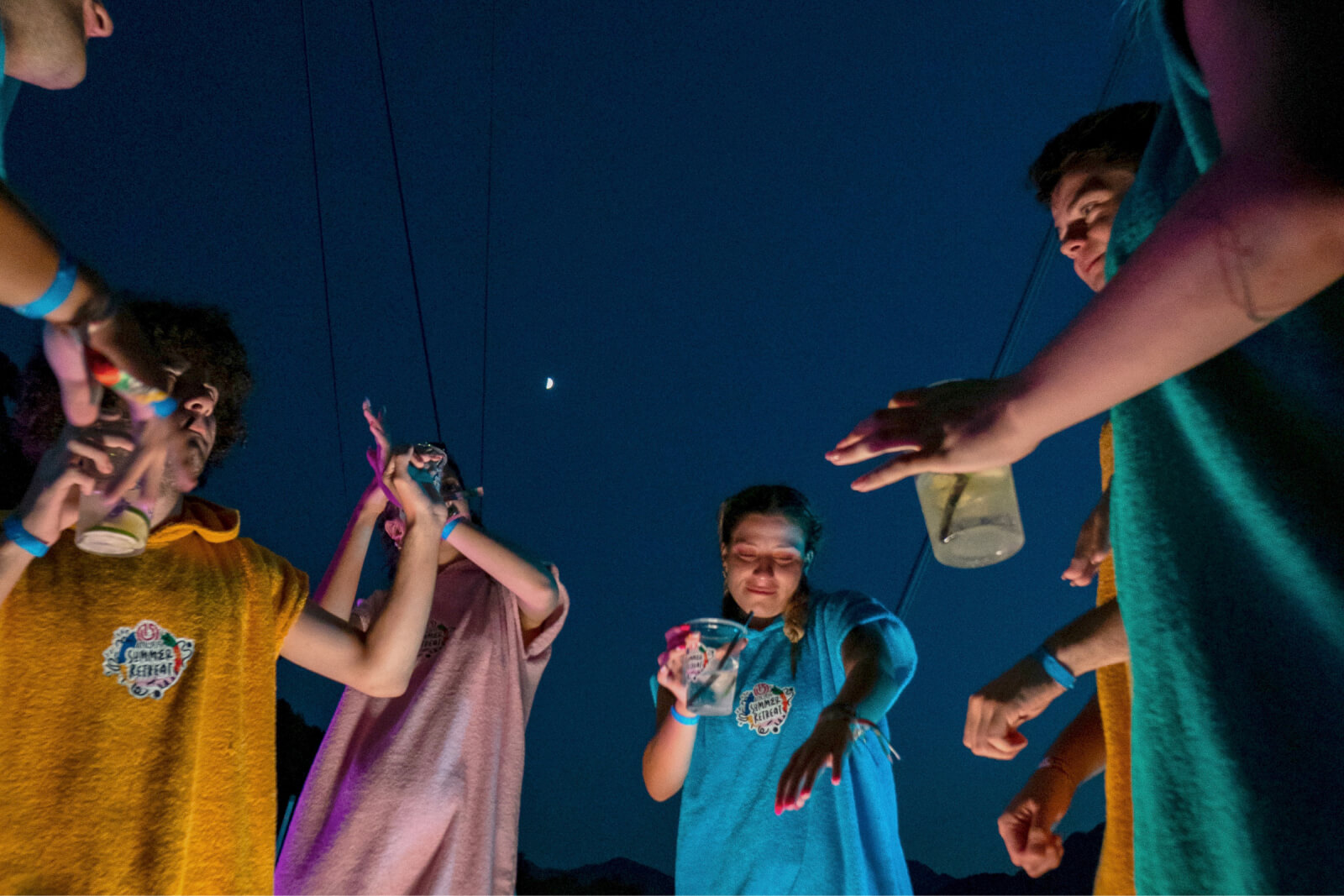 A group of young people dancing in orange, pink, blue, and green towel beach ponchos and holding plastic cups at night.