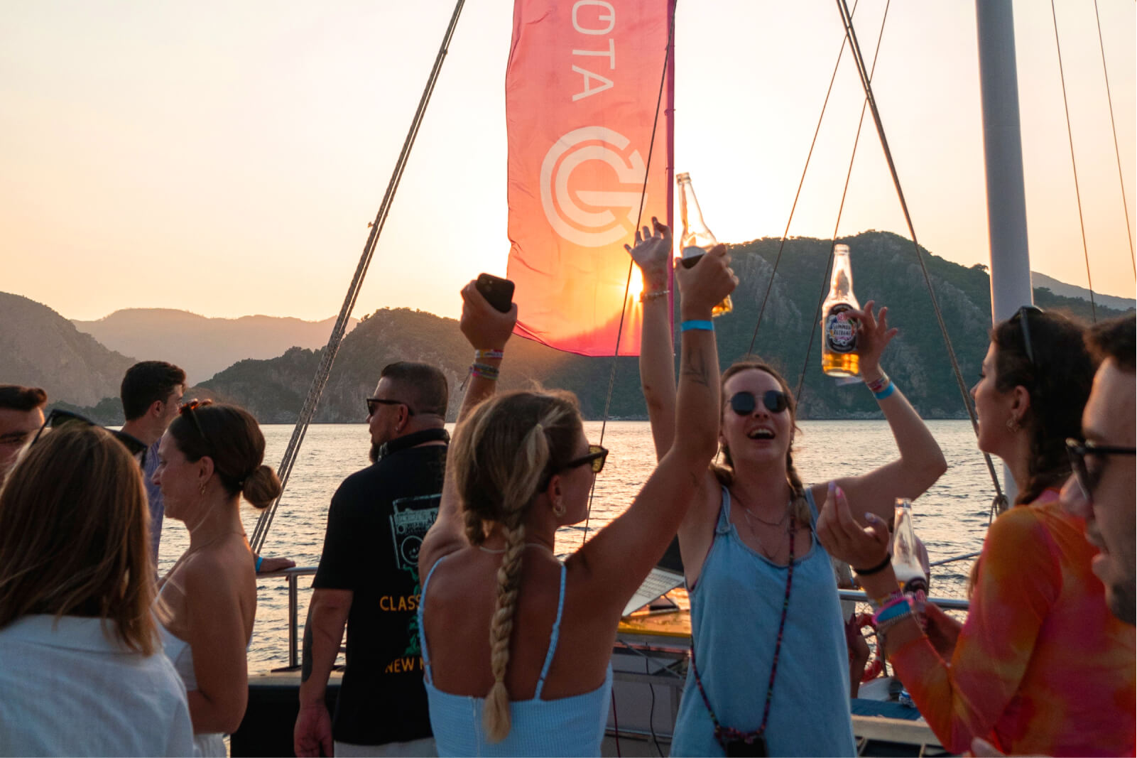 A group of people and two blond girls raising their beer in the air by the sea, and the Atolye15 logo on a red flag.