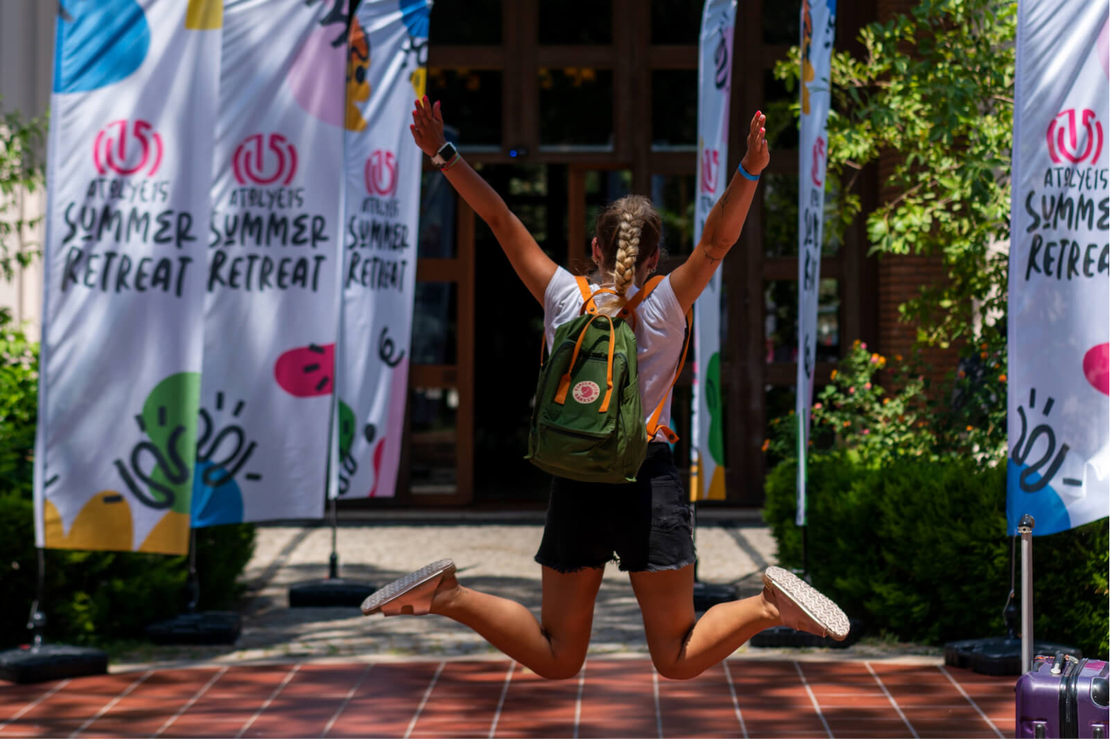 A blonde girl with braids, wearing denim shorts and a white t-shirt, jumping in the air next to Atolye15 Summer Retreat banners.
