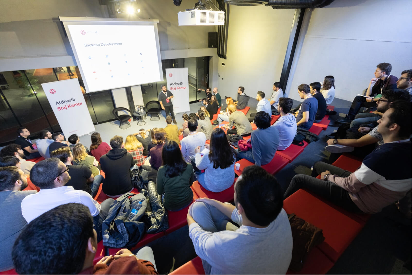 People at a lecture hall listening to a man’s presentation on backend development in front of “Atölye15 Staj Kampı” banners.
