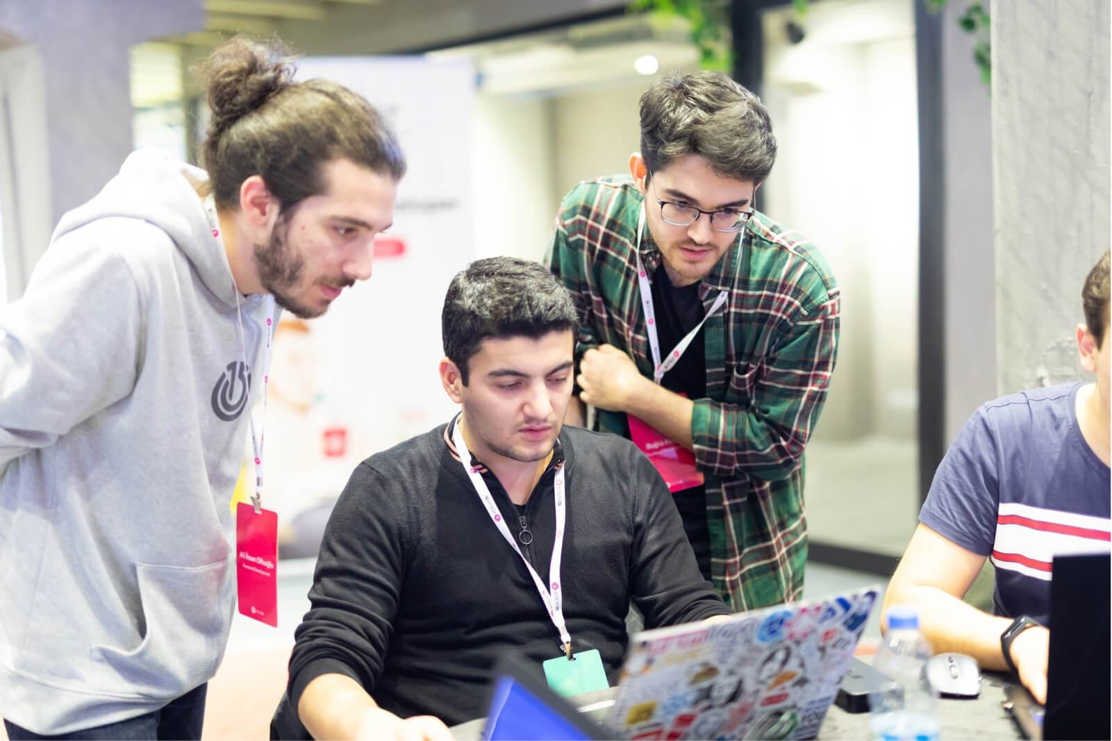 A man with a bun in a grey Atolye15 hoodie and another man in a green plaid shirt watching over a man working on his laptop.
