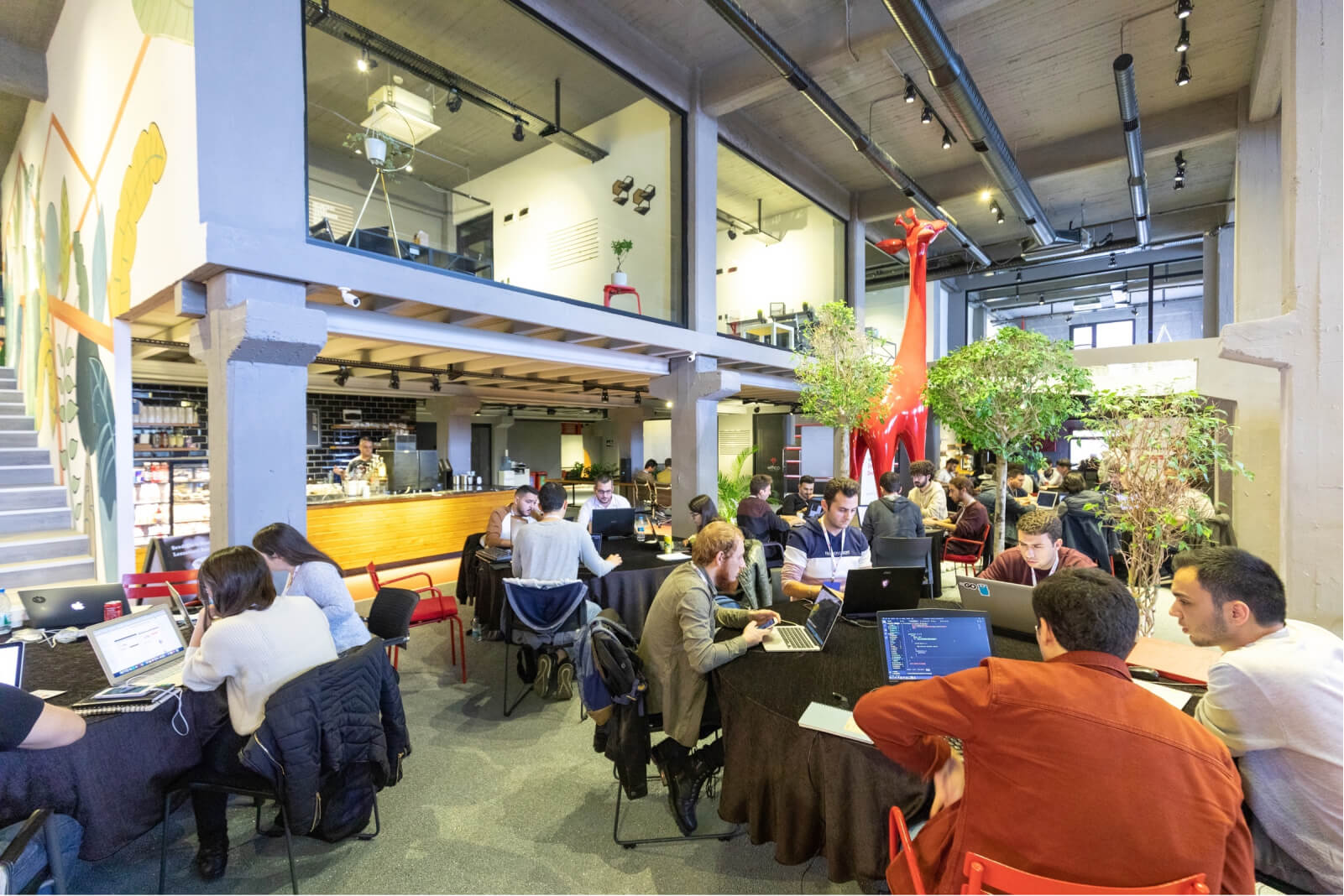 An interior space filled with tables of Bootcamp attendees working on their computers, plants, and a big red giraffe statue.