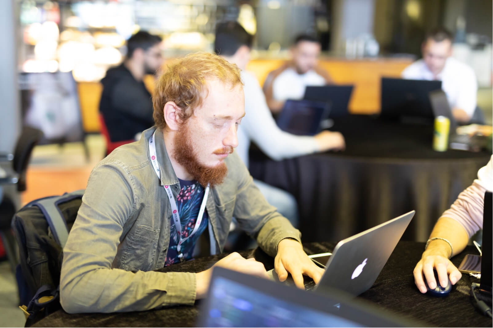 A ginger man with a beard wearing a green shirt and working on his space grey MacBook next to another table of men.