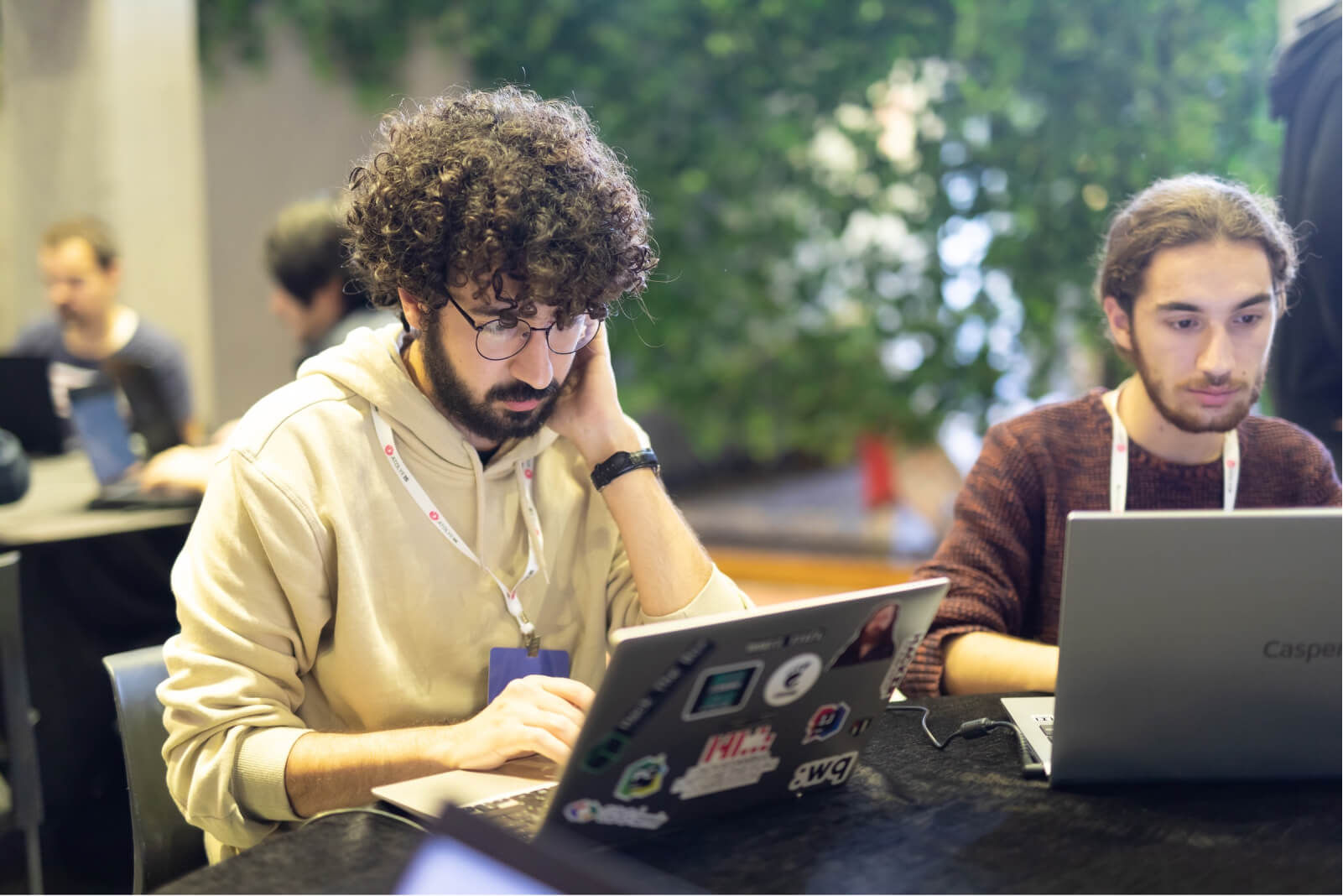 A curly-haired man wearing glasses and a hoodie working on a laptop covered with stickers, sitting next to a long-haired man.