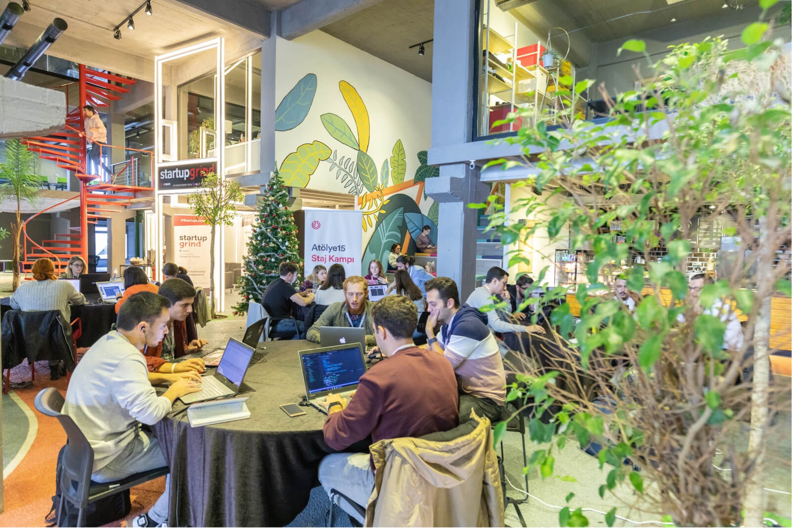 People working on their laptops at round tables in a place filled with plants and a banner saying “Atölye15 Staj Kampı.”