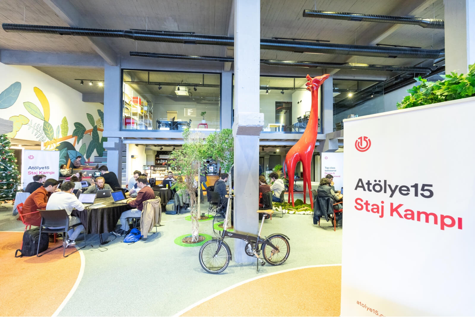The interior of a building where people are working on their laptops at round tables and a banner that says “Atölye15 Staj Kampı.”