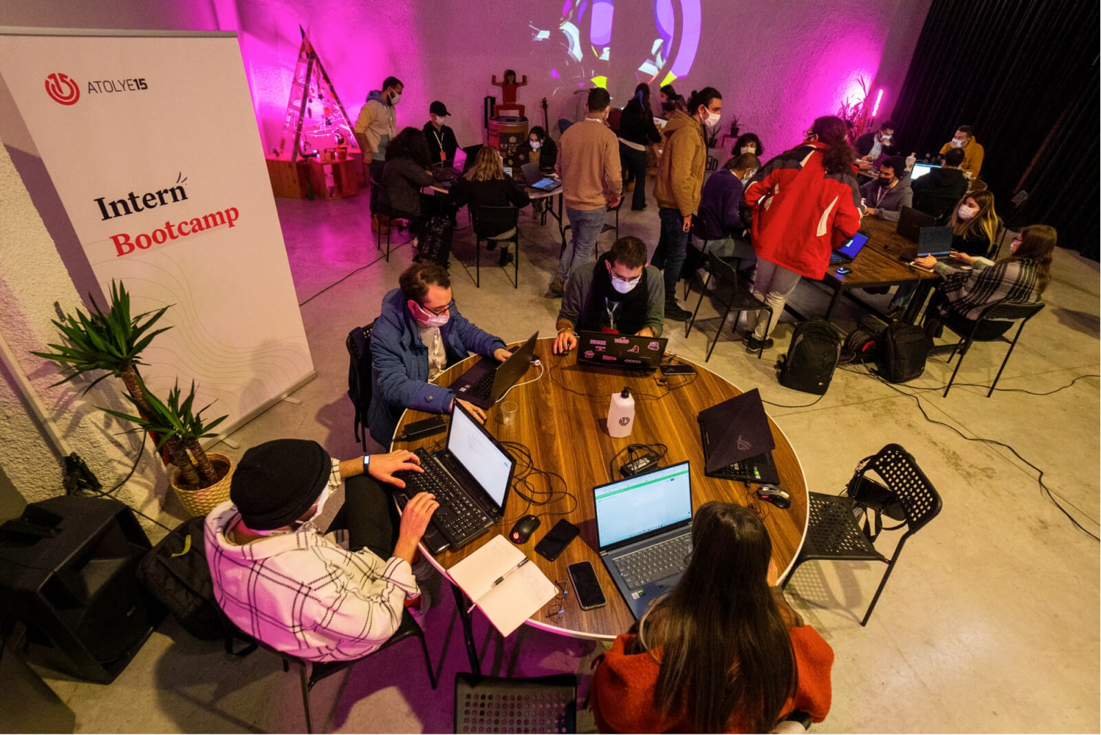 A group of people, sitting around a round table working on their laptops and a banner that says “Atolye15 Intern Bootcamp.”