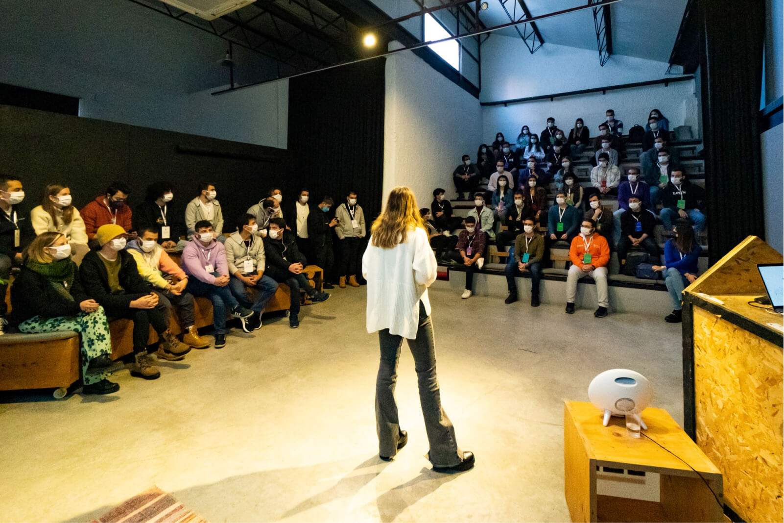 The back of a woman in a white sweater giving a presentation in front of a crowd of people wearing face masks.